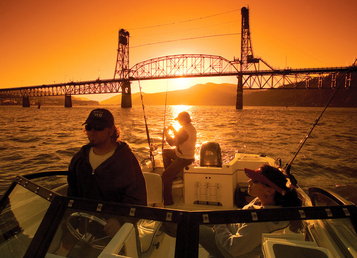 Against the backdrop of a colorful setting sun and a bridge, three crew members in their fishing boat cruise back to the dock.